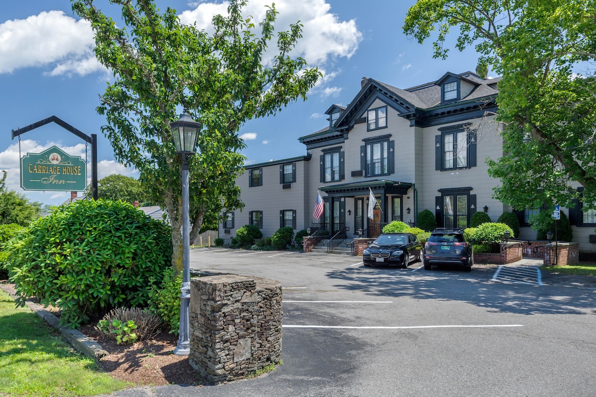 The exterior of the Carriage House is showcased with a well-maintained façade featuring classic architecture. A spacious parking area is visible in the foreground, accommodating multiple vehicles. Lush greenery surrounds the building, and an American flag is displayed on the entrance.