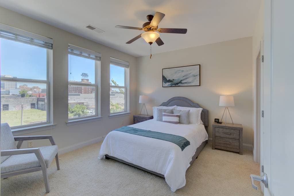 A well-lit bedroom features a spacious bed with a light-colored duvet and decorative pillows. Two bedside tables with lamps are positioned beside the bed. A comfortable chair is placed next to a window, revealing views of downtown Memphis.