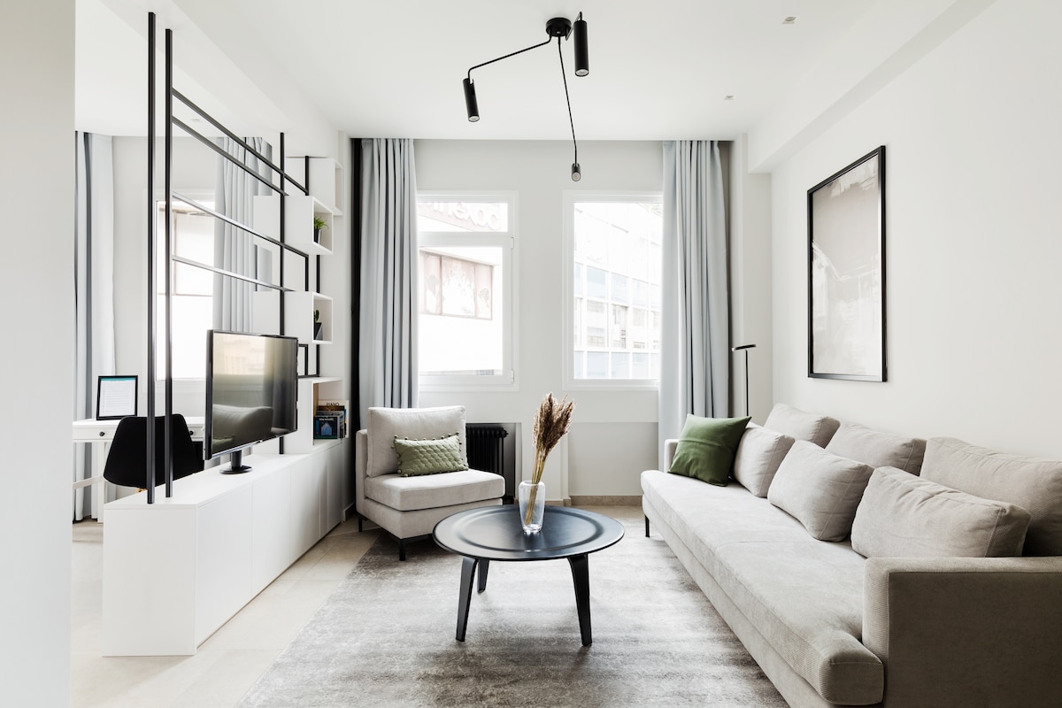 A modern living area is highlighted by a large gray sofa and a cozy armchair. A circular coffee table sits on a soft rug, while a television is mounted on a white media console. Natural light filters through two windows, enhancing the open atmosphere.