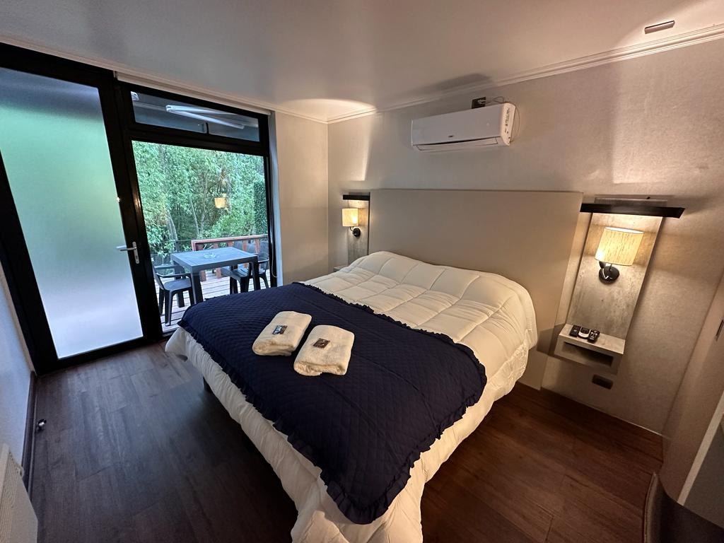 A bright bedroom features a spacious bed adorned with a navy blue quilt and neatly arranged towels. Soft lighting from bedside sconces enhances the calming atmosphere. Glass doors provide a view of the outdoor seating area surrounded by greenery.