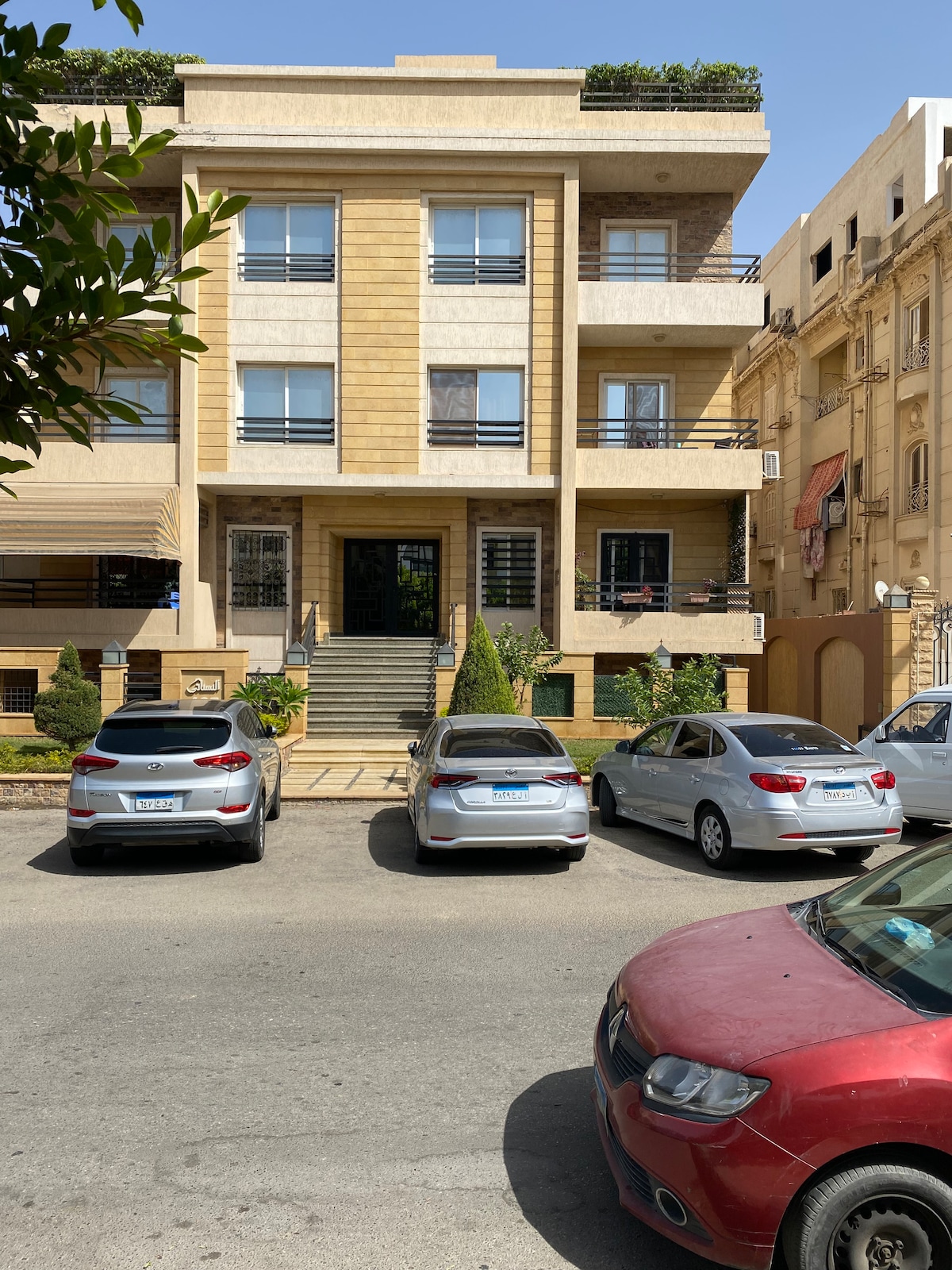 The building's exterior features a symmetrical design with a golden stone façade and multiple balconies. A staircase leads to the main entrance, framed by neatly arranged shrubs and small trees. Several parked cars line the street, complementing the urban environment.