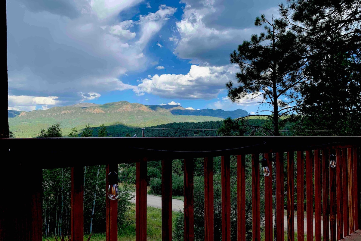 The outdoor view reveals expansive mountain scenery under a partly cloudy sky, with lush greenery in the foreground. A wooden railing frames the image, enhancing the sense of openness and connection to nature.