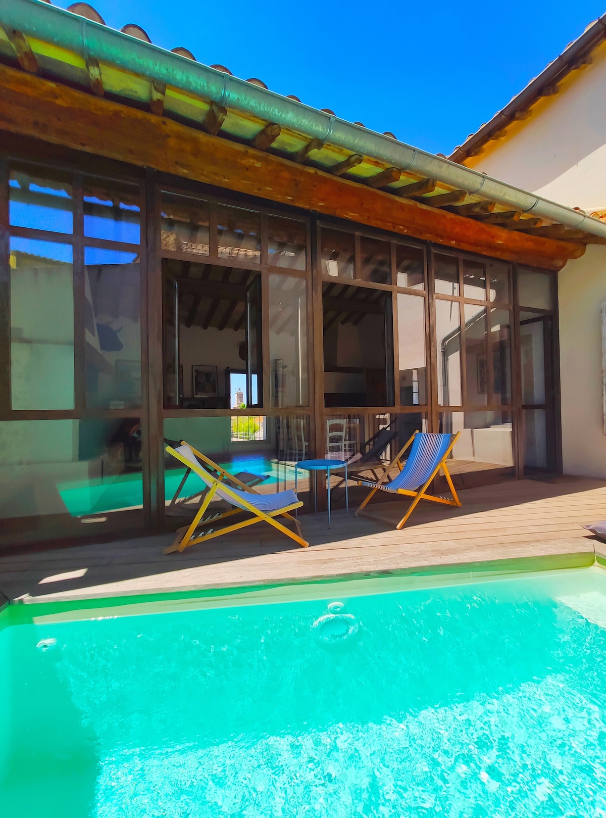 A terrace area features two lounge chairs positioned beside a sparkling pool, with large glass doors providing a view of the interior. The space is illuminated by natural light, highlighting the wooden ceiling beams and creating an open, airy ambiance.