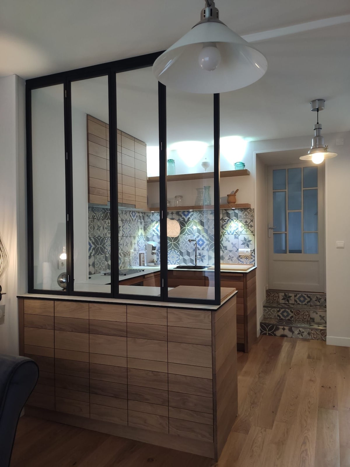 A modern kitchen area is visible, featuring light wood cabinetry and intricate tiled backsplash. Soft lighting highlights the workspace, and a combination of glass panels and dark frames separates it from the living space. A staircase is visible in the background, leading to another area.