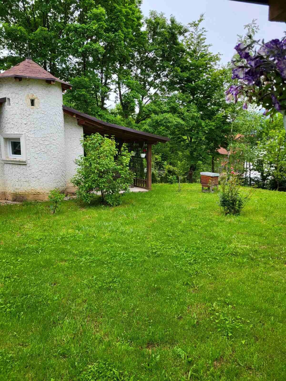 A charming villa exterior is surrounded by lush greenery. A small garden area features well-maintained grass and young trees, with the villa’s textured white wall visible. A covered porch provides shaded seating, enhancing relaxation in a serene natural setting.