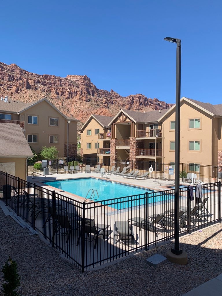 The image displays a clear view of a swimming pool surrounded by a secure black fence, with lounge chairs arranged neatly around it. In the background, a red rock formation rises against the bright blue sky, while adjacent buildings offer a warm, welcoming appearance.
