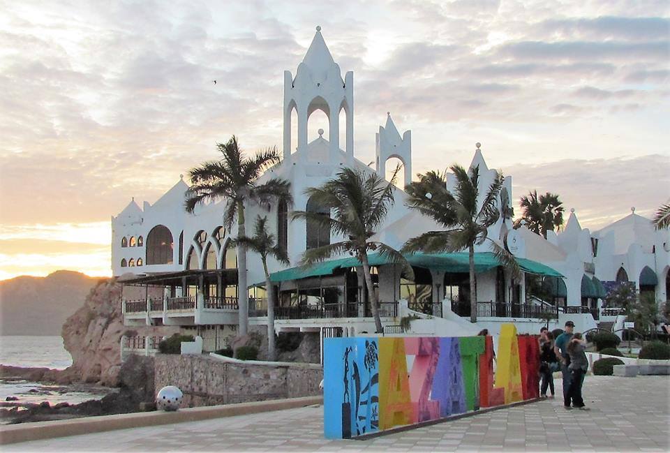 An expansive beachfront structure with a distinctive architectural style is visible, featuring white walls and arched windows. Palm trees sway gently in the foreground, with a colorful mural displayed on a nearby wall. A serene sunset casts warm tones across the sky and water.
