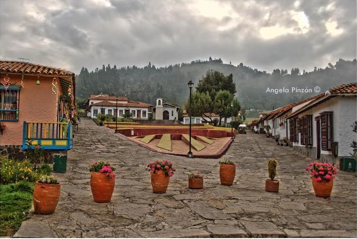 A cobblestone pathway is framed by terracotta pots filled with colorful flowers. Rustic buildings with traditional architecture are visible in the background, surrounded by lush green mountains under a cloudy sky, creating a serene environment.