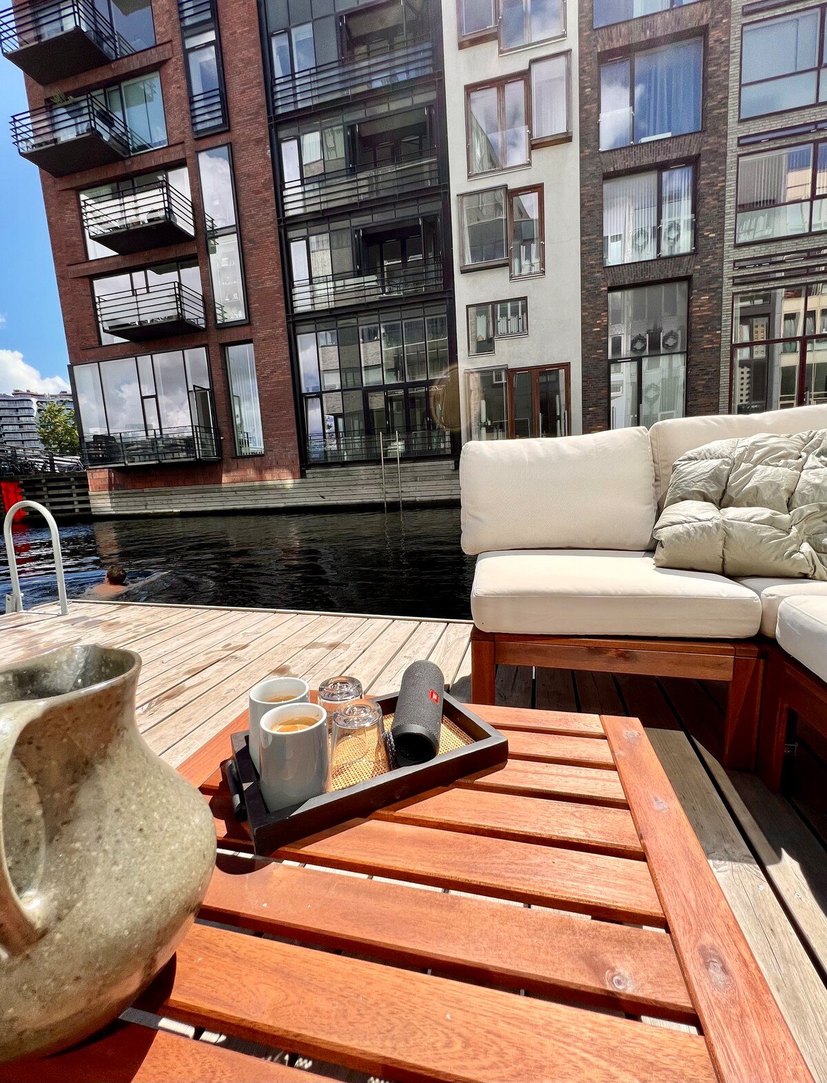 A private floating terrace is depicted, featuring a wooden table with a tray holding two cups and sugar containers. A comfortable seating area with light-colored cushions overlooks the tranquil water, while modern apartment buildings are visible in the background, reflecting sunlight.