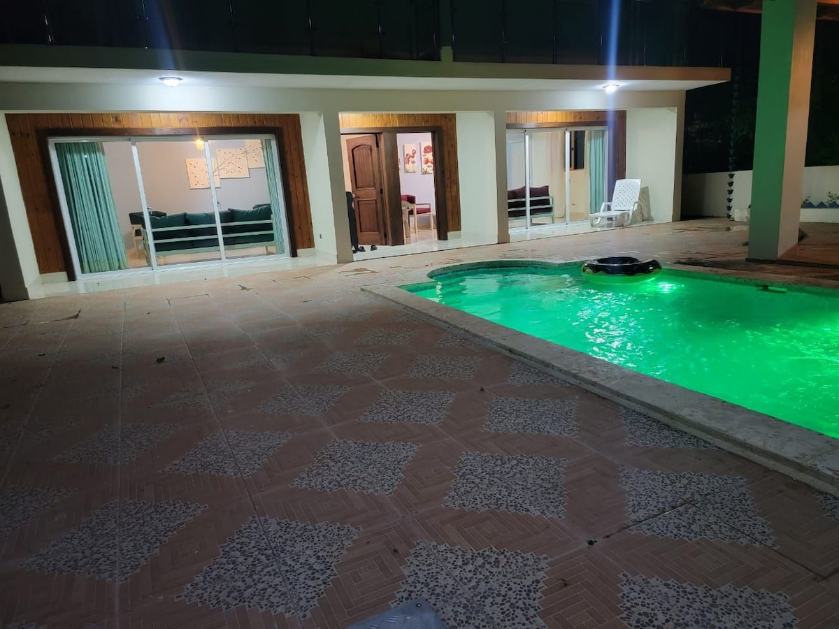 An outdoor space features a swimming pool with illuminated green water, surrounded by a patterned tile patio. Large glass doors provide a view of the living area, showcasing a sofa and additional seating. Soft overhead lighting helps to create a welcoming ambiance.