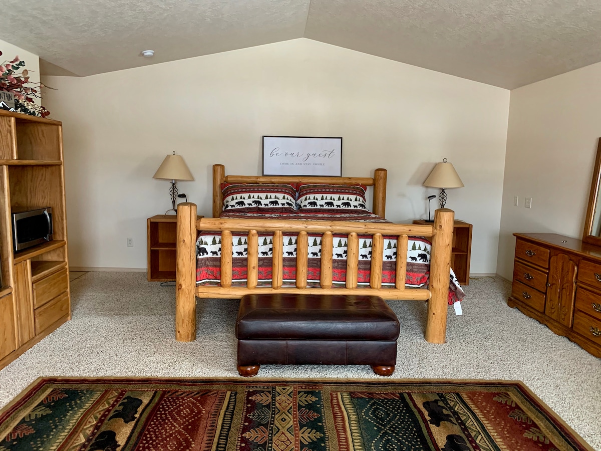 The spacious master suite includes a large wooden bed with a patterned blanket, accompanied by two bedside lamps. A dark ottoman is positioned at the foot of the bed. Wooden furniture lines the walls, enhancing the rustic charm of the room.