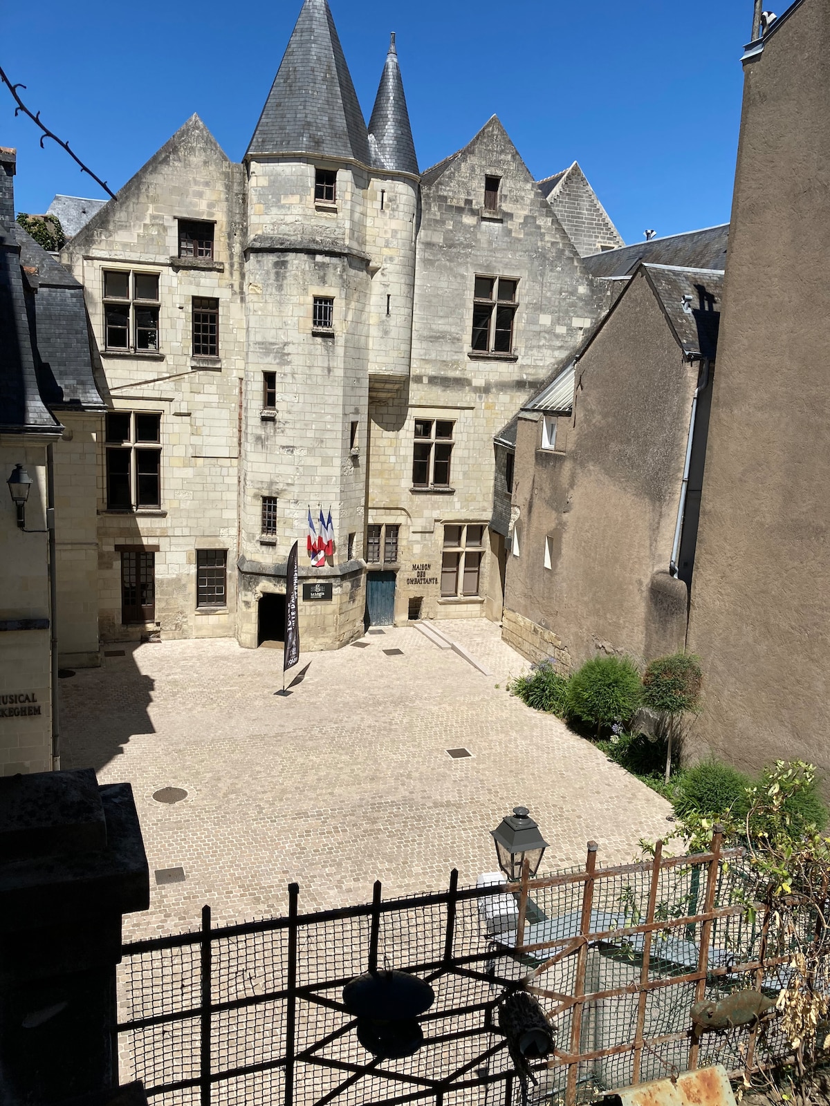 A historic stone courtyard is visible, featuring tall, turreted buildings that showcase architectural details. The cobblestone surface is surrounded by greenery, and a lantern stands at the edge. The clear blue sky adds brightness to the scene, highlighting the vibrant facade of the structures.