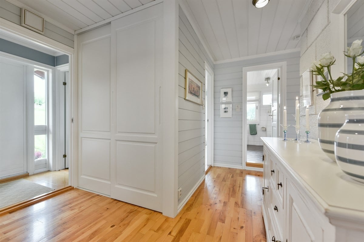 A bright hallway is presented with white wooden paneling and warm wooden flooring. Double doors provide storage space on one side, while stylish decor and a vase of flowers are positioned on a console table. Doorways lead to other rooms, enhancing the home’s spacious feel.
