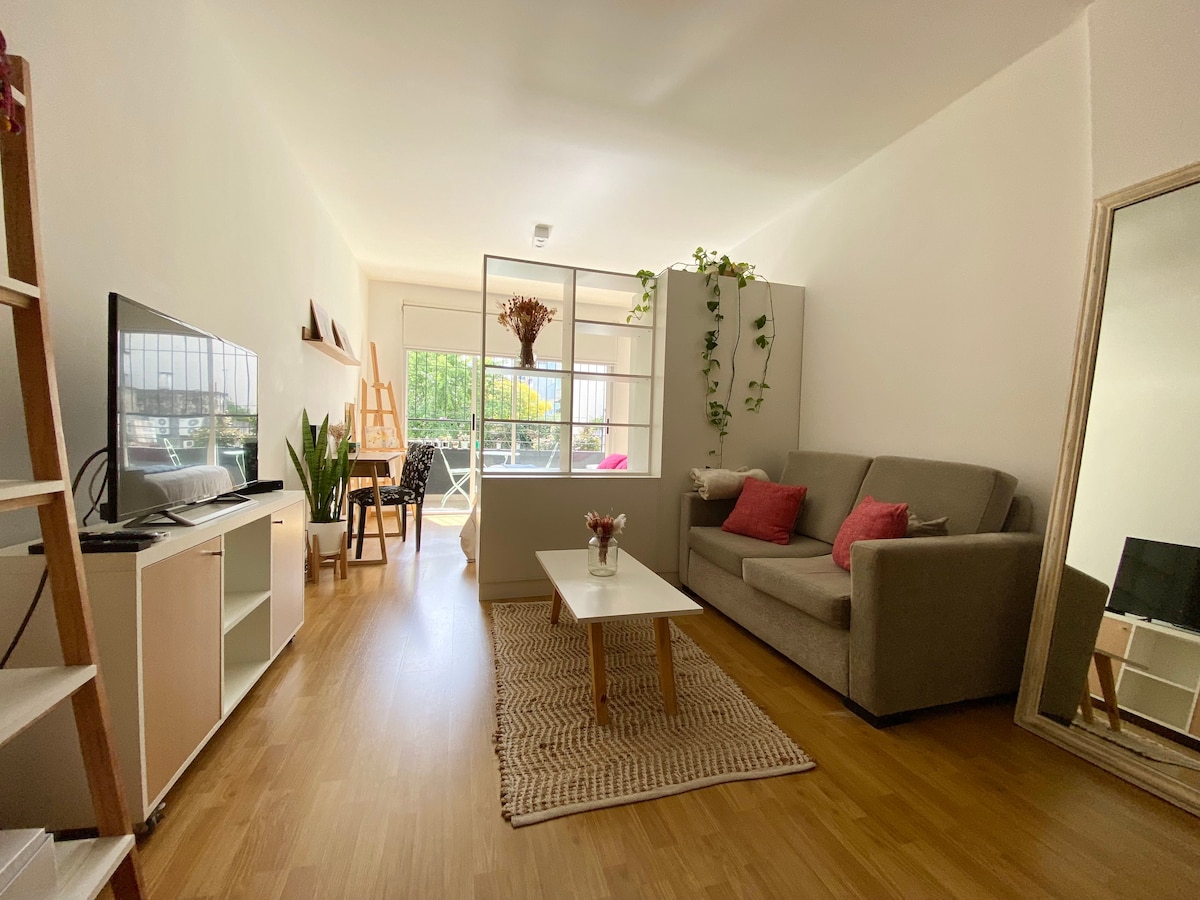 A bright living area features a comfortable gray sofa with decorative pillows, a wooden coffee table, and a decorative rug. A large window allows natural light to fill the space, revealing a glimpse of greenery outside. A tall mirror and shelving unit are present, enhancing the room's openness.