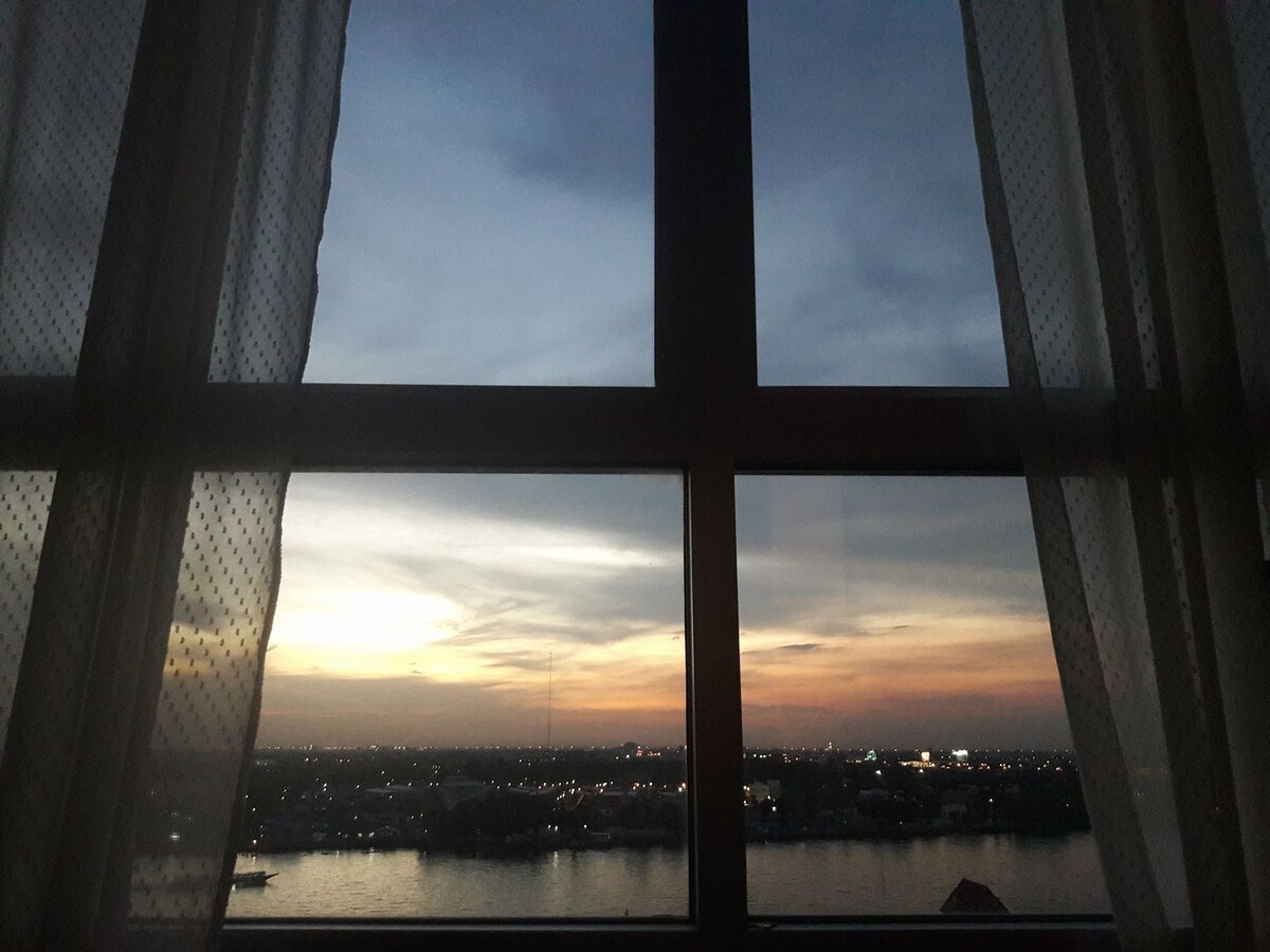 A large window frames a panoramic view of the river at sunset, with soft hues of orange and blue blending in the sky. Light curtains gently drape on either side, allowing natural light to filter into the space while highlighting the serene evening scene.