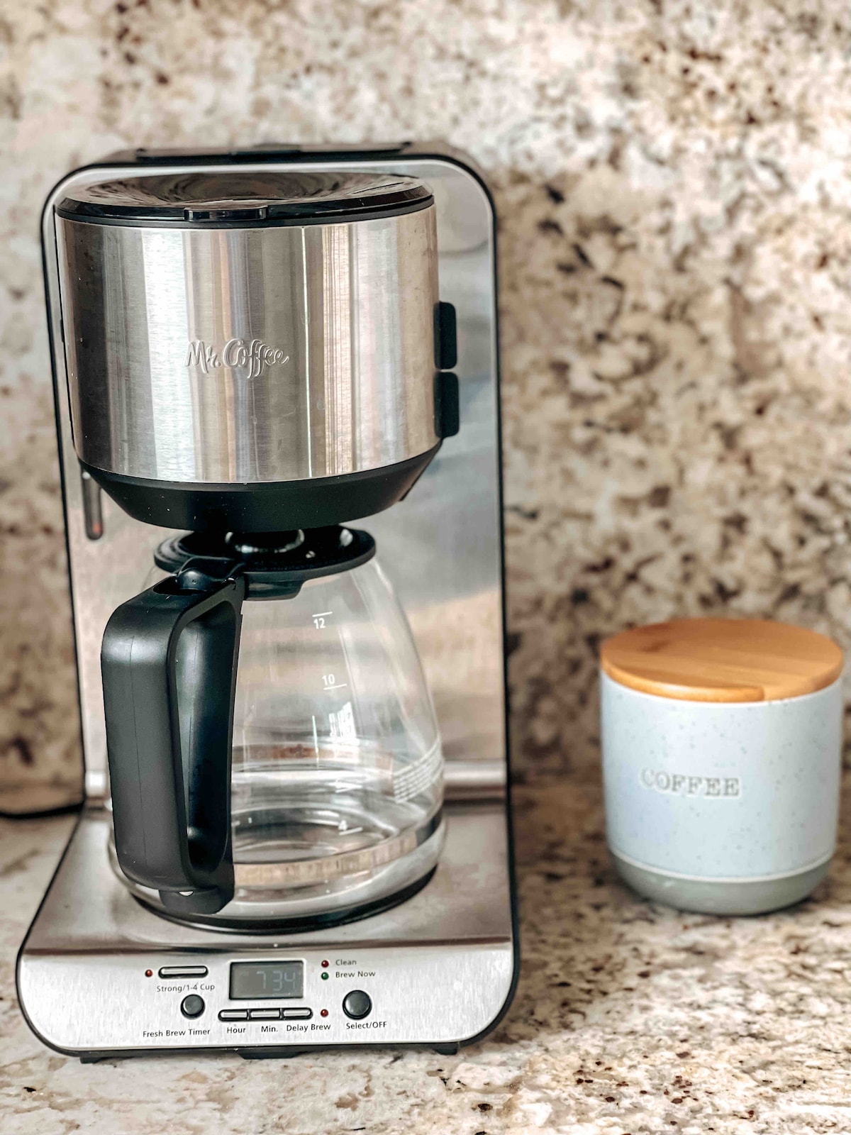 A coffee maker with a stainless steel finish is positioned on a polished granite countertop. A glass carafe sits underneath, marked with measurement indicators. Nearby, a light blue ceramic container with a wooden lid is labeled 'coffee', providing an organized space for coffee essentials.