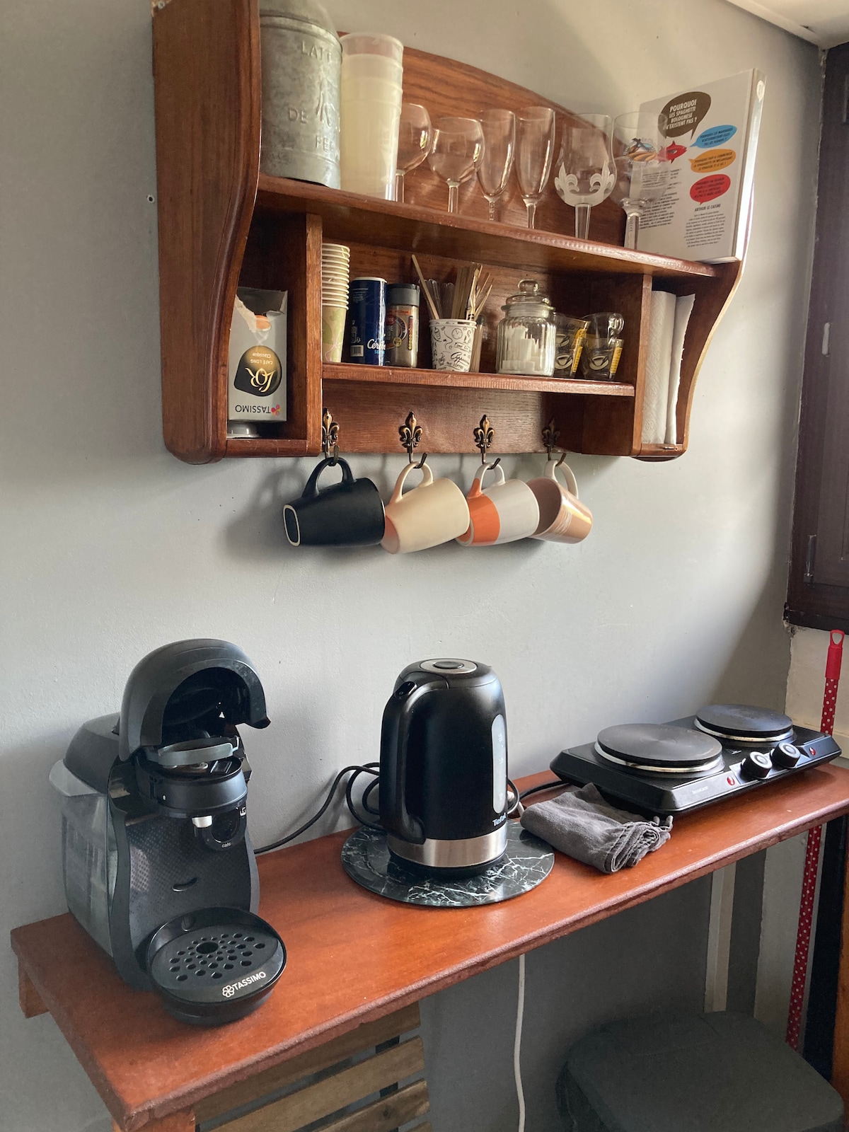 A well-organized kitchenette area features a wooden shelf displaying glasses, cups, and condiments. A coffee machine, electric kettle, and cooking appliances are arranged on a countertop, with two tea towels positioned neatly. The wall is painted in soft tones, enhancing the functional space.