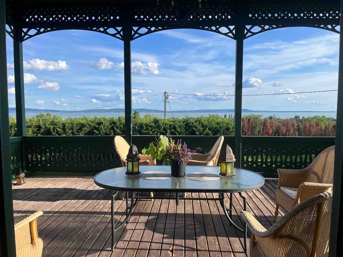 A spacious terrace is presented, featuring a large table surrounded by several comfortable chairs. Decorative lanterns and potted plants are placed on the table. A panoramic view of rolling hills and a clear sky is visible, enhancing the outdoor ambiance.