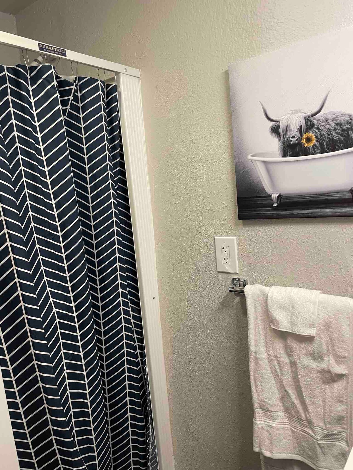 A bathroom area is depicted featuring a patterned shower curtain in navy and white. A decorative piece displaying a cow in a bathtub is mounted on the wall. A towel is neatly hung on a wall-mounted rack beside the shower.
