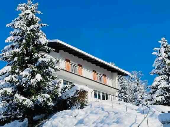 The house is set against a clear blue sky, surrounded by snow-covered trees. A gently sloping pathway leads to the entrance, with snow layered on the ground and rooftops. Balconies with wooden railings offer a charming contrast to the pristine white landscape.