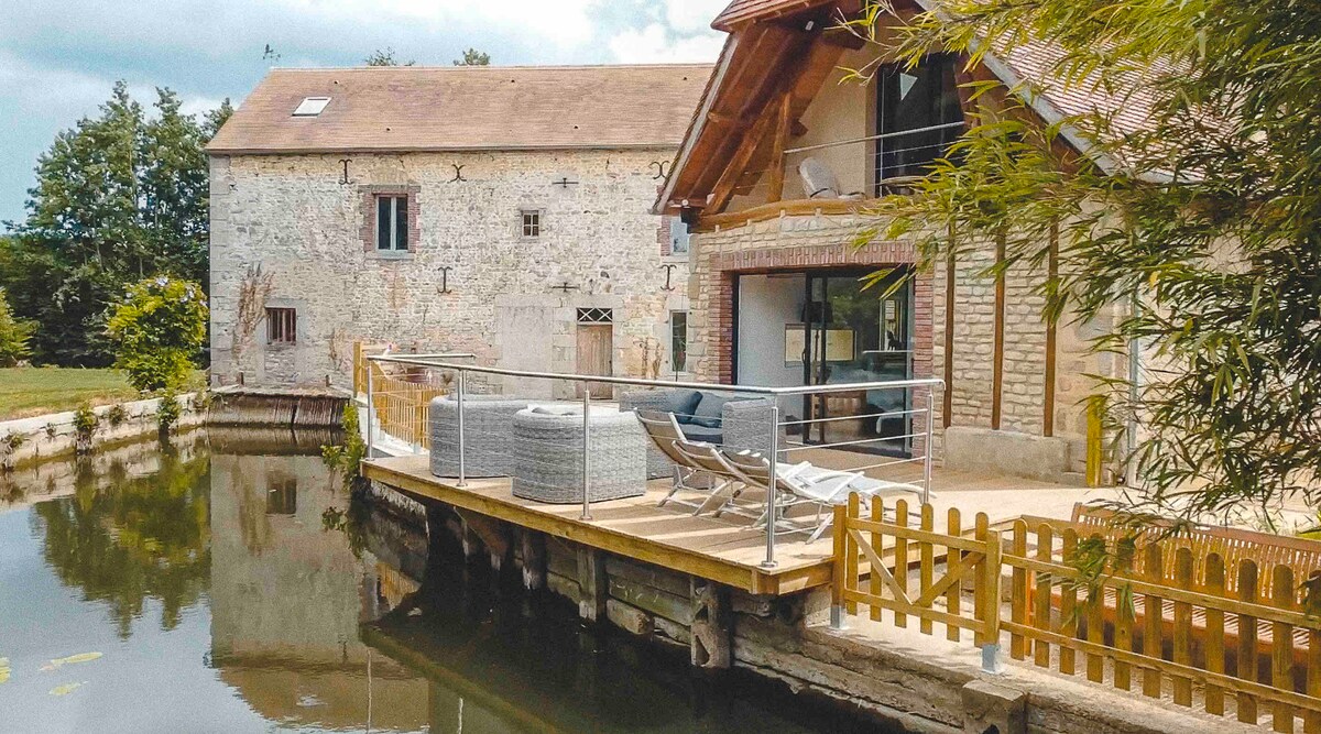 A charming outdoor terrace extends over the water, featuring a space for seating complemented by sun loungers. A low wooden fence provides a clear boundary, while the rustic stone building reflects the serene surroundings of lush greenery and a calm waterway.