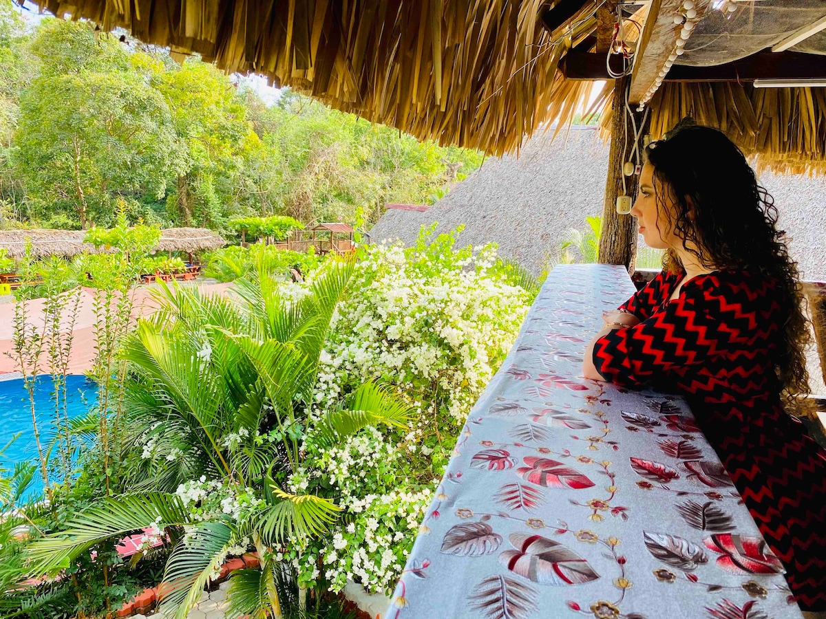 A view from a wooden terrace showcases lush greenery and vibrant flowers surrounding a swimming pool. The scene is enhanced by the rustic thatched roof and the tranquil atmosphere created by the natural landscape.