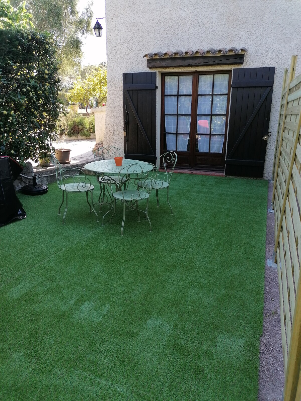 A private terrace features artificial grass, offering a space for outdoor meals and relaxation. A round table is surrounded by four metal chairs. Double doors with wooden shutters lead into the studio, framed by greenery.