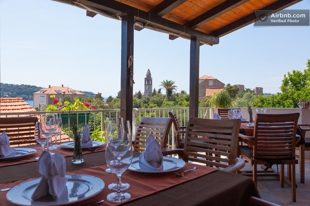 An outdoor dining area is presented with wooden tables set for a meal. Elegant tableware and neatly folded napkins are visible. The surrounding greenery and rooftops create a scenic backdrop, with a distant clock tower adding to the charming view.