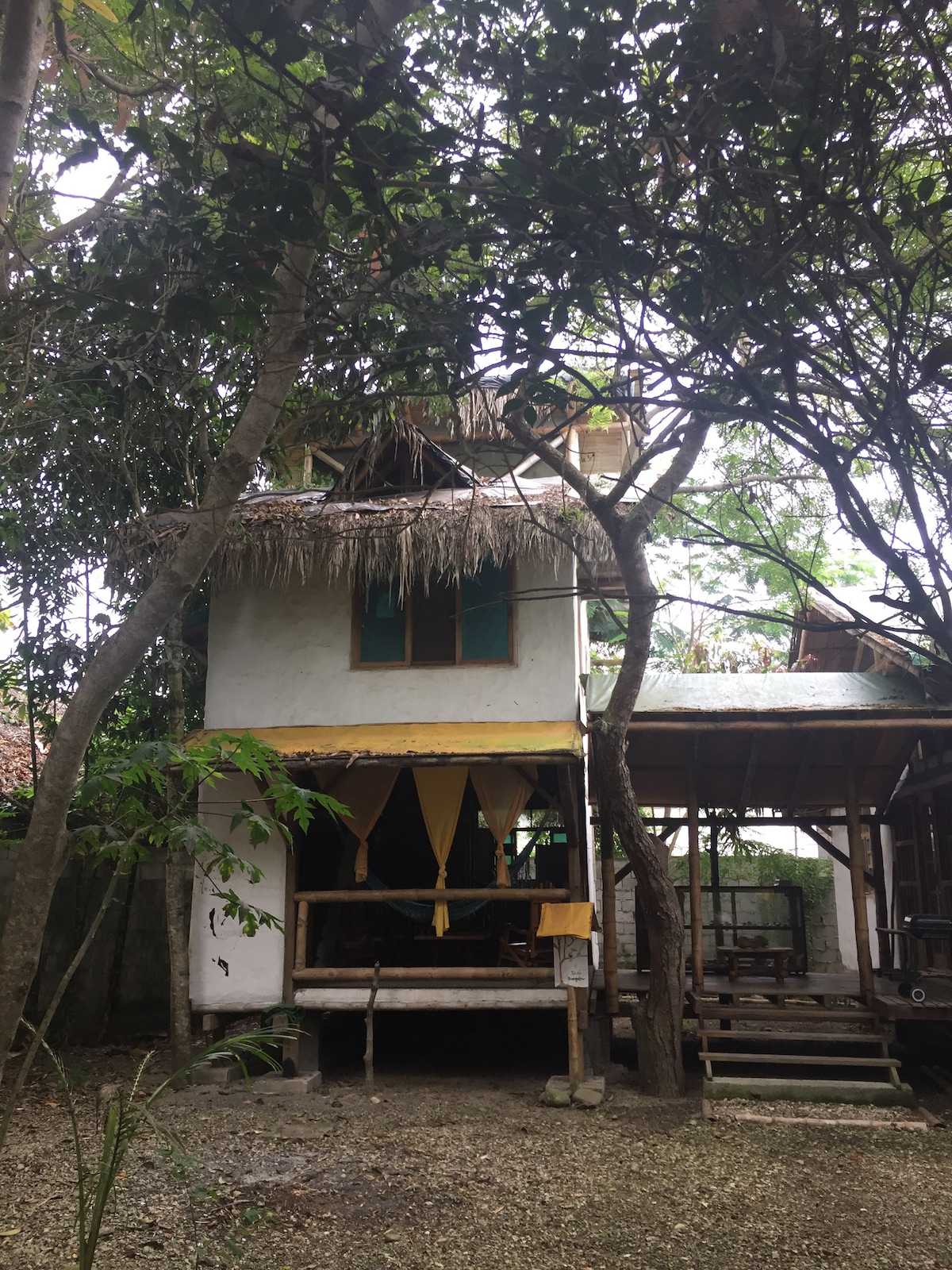 The three-story bungalow features a thatched roof and is surrounded by lush vegetation. A covered porch is visible at the front, with yellow drapes that softly frame the entrance. The structure blends harmoniously with the natural environment, providing a secluded and inviting atmosphere.