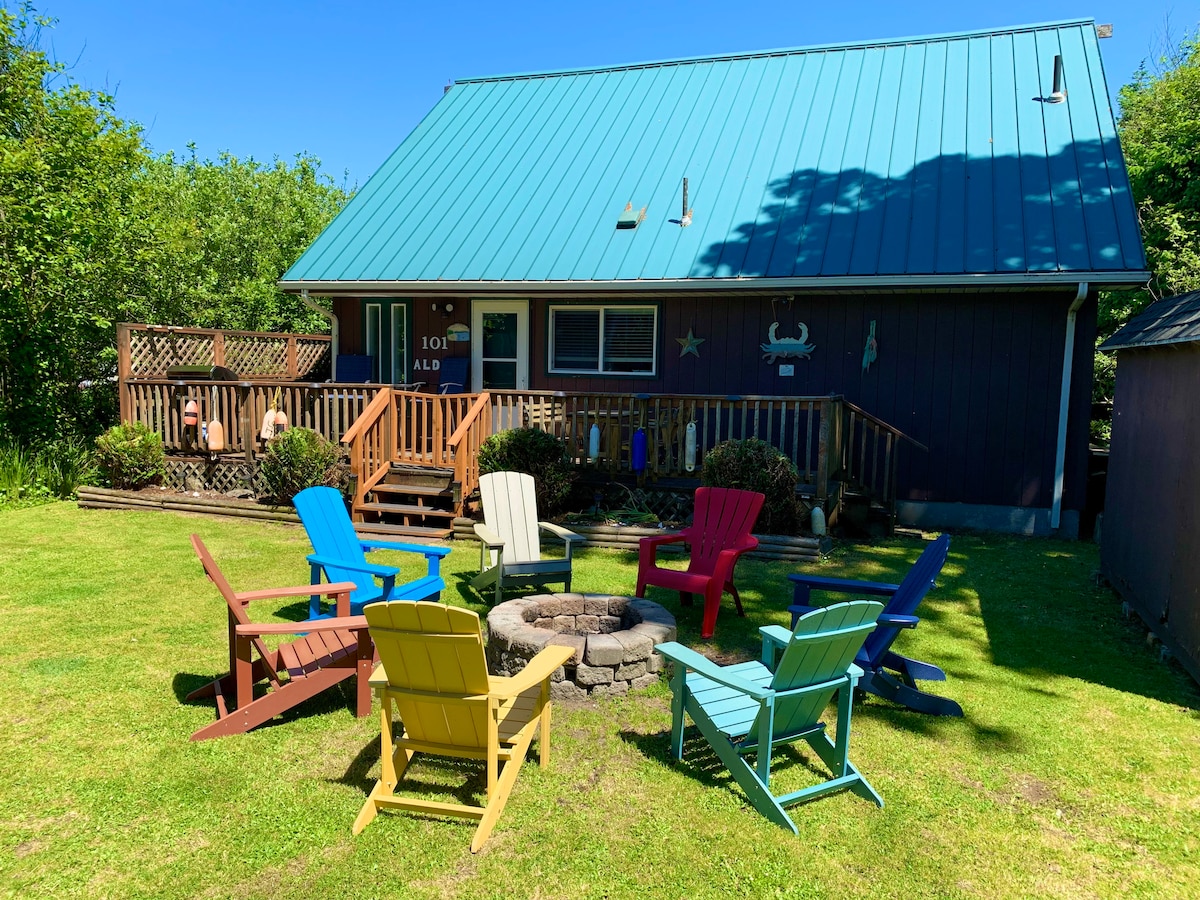 A fire pit surrounded by colorful Adirondack chairs is set in a spacious grassy yard. The cabin features a blue metal roof and multiple wooden deck areas, providing inviting spaces for relaxation and gatherings. Lush greenery frames the property, enhancing the natural setting.
