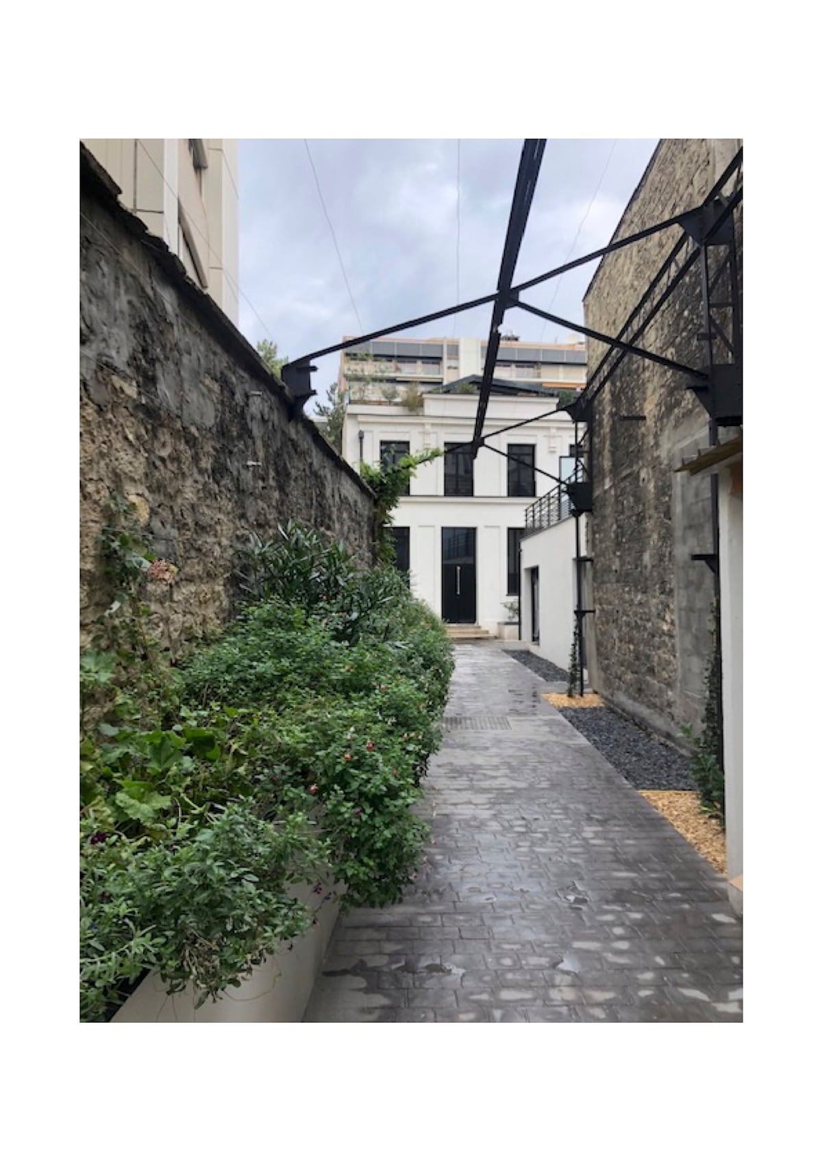 A narrow path is framed by low stone walls, lined with lush greenery and potted plants. The walkway is made of cobblestones, leading towards a white building with dark doors and large windows, accented by exposed metal beams above.