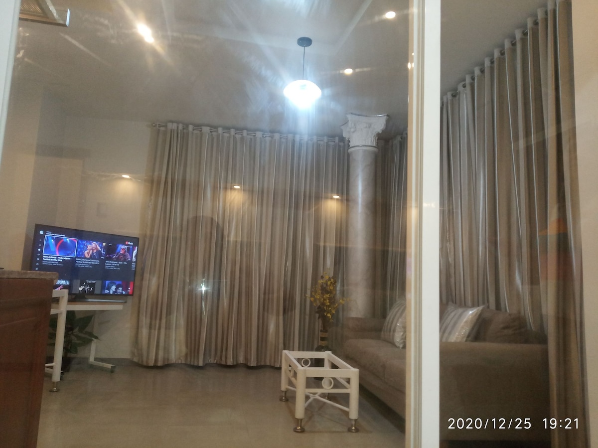 A living area is visible featuring a comfortable couch next to a decorative column. A coffee table is positioned in front of the couch. Soft lighting from a ceiling fixture and wall-mounted lights creates a relaxed ambiance, complemented by sheer curtains framing a large window.