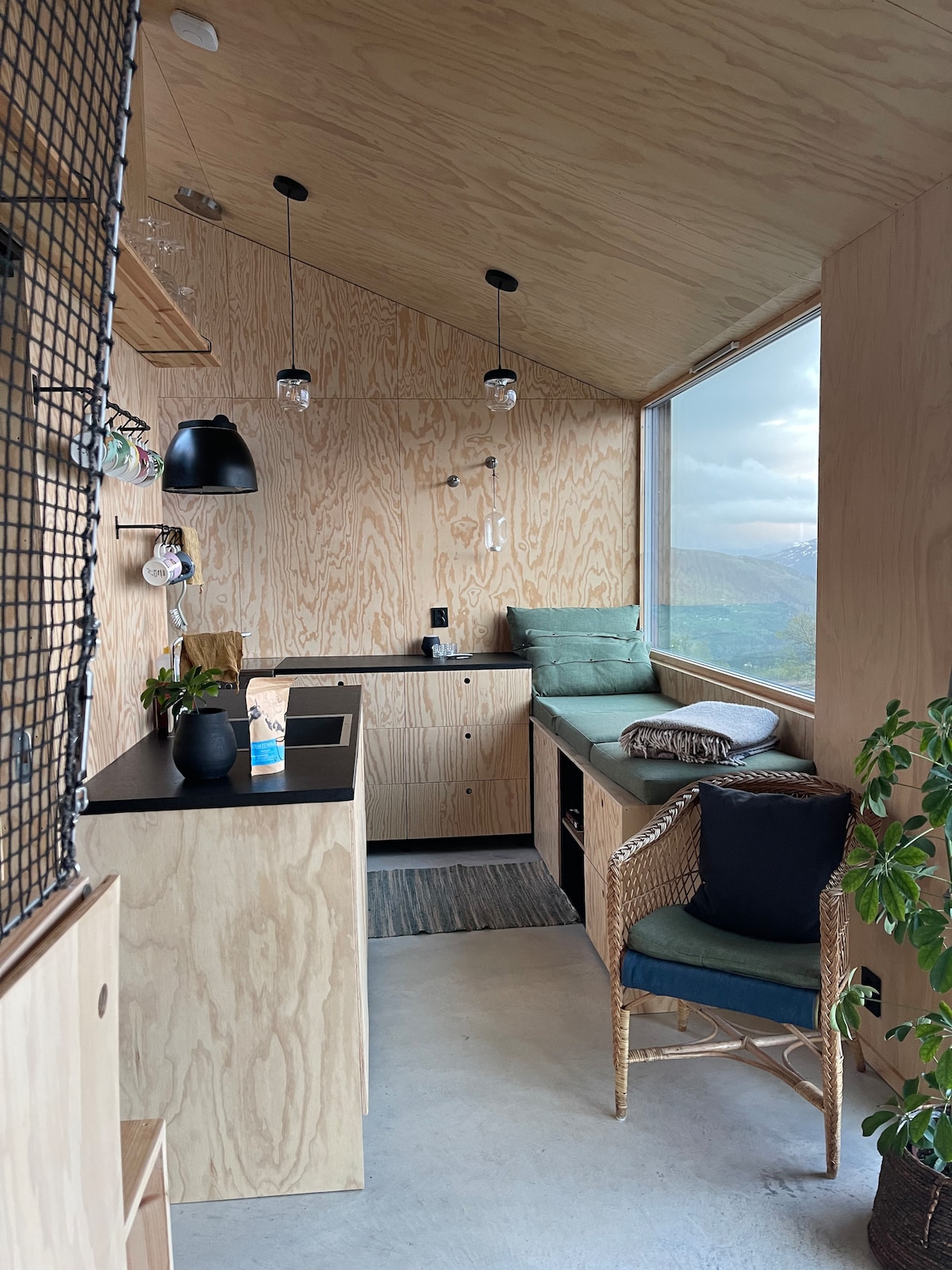 A modern interior space is highlighted by warm wooden walls and sleek furnishings. A small kitchenette features minimalistic cabinetry, while a comfortable seating area is positioned by large windows, allowing for expansive views. A green armchair and decorative plants add to the inviting ambiance.