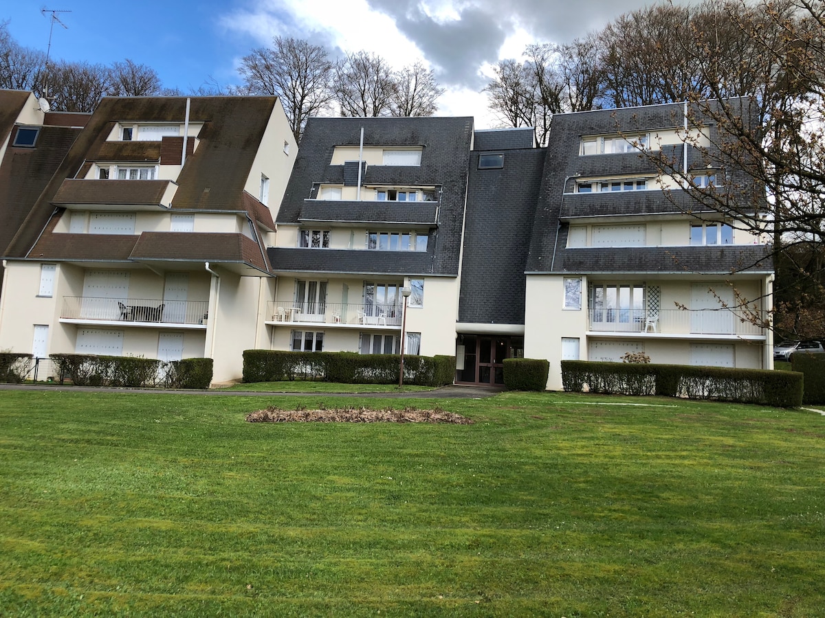 An elegant multi-story building is set in a landscaped area featuring neatly trimmed grass and shrubbery. Spacious balconies are visible on each level, with windows allowing natural light into the interiors. The surrounding trees add a touch of greenery to the peaceful environment.