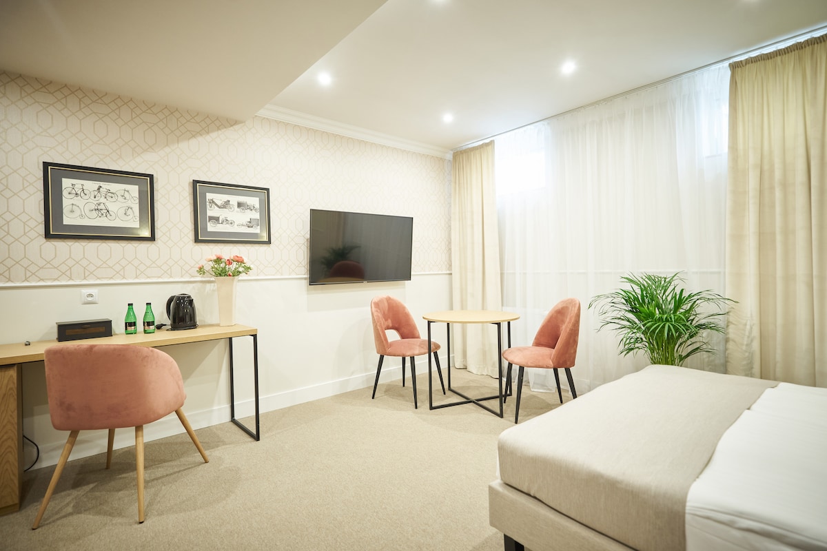 A modern room features a spacious bed and a comfortable seating area with two pink chairs and a round table. A wooden desk with a kettle and complimentary bottled water is visible, alongside a wall-mounted television. Natural light filters through sheer curtains.