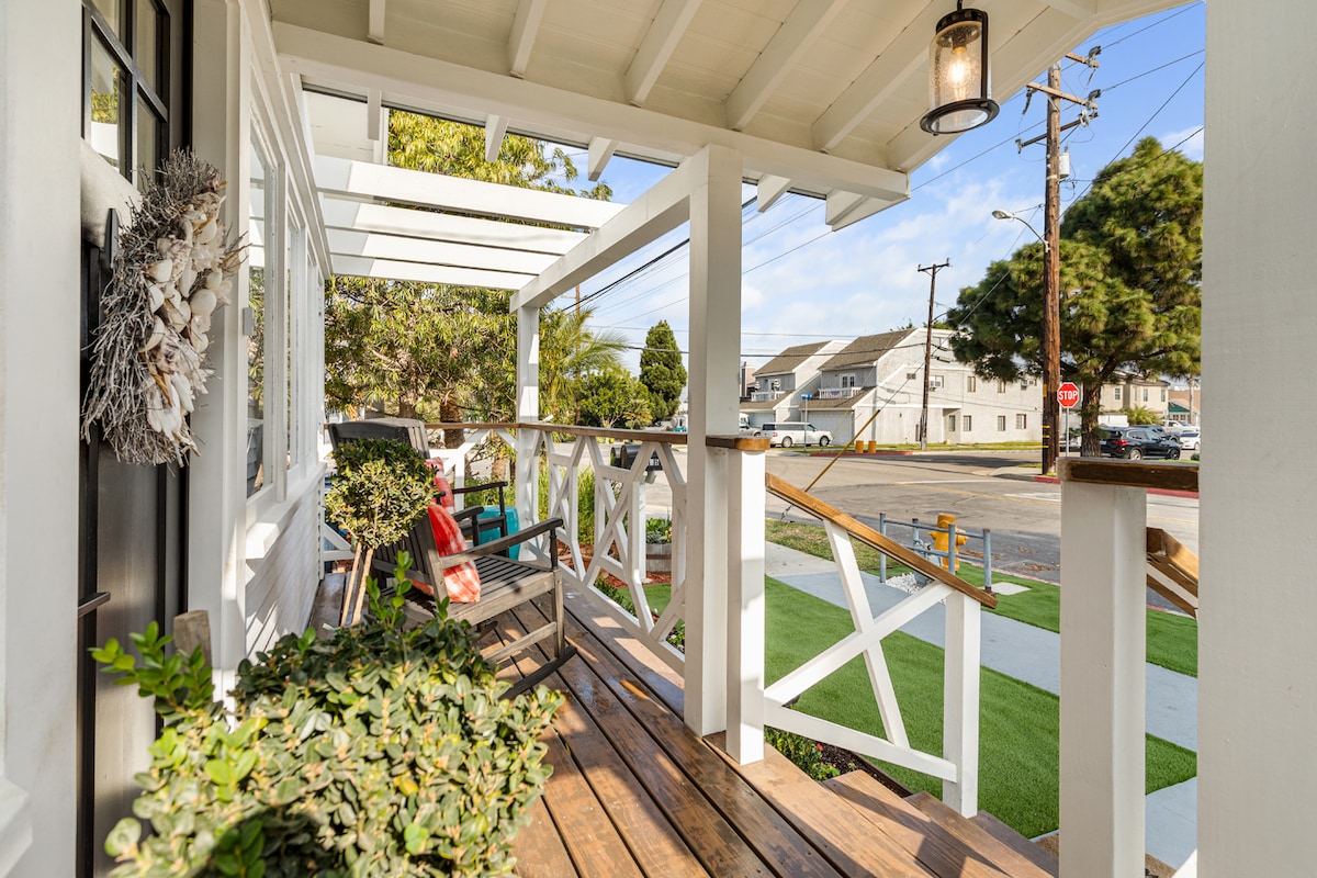The exterior deck is framed by a white railing, featuring comfortable seating and greenery. Natural light illuminates the wooden floor, while the surrounding area reveals a well-maintained lawn and the quiet street beyond.
