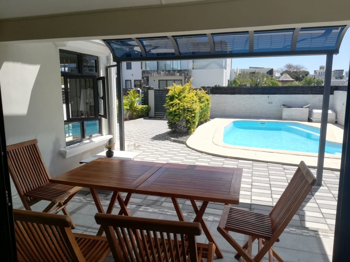 An outdoor dining area is shown, featuring a wooden table surrounded by several chairs. The space is shaded by a canopy, overlooking a private pool area with surrounding greenery and paving. The pool is visible in the background, adding a serene element to the setting.