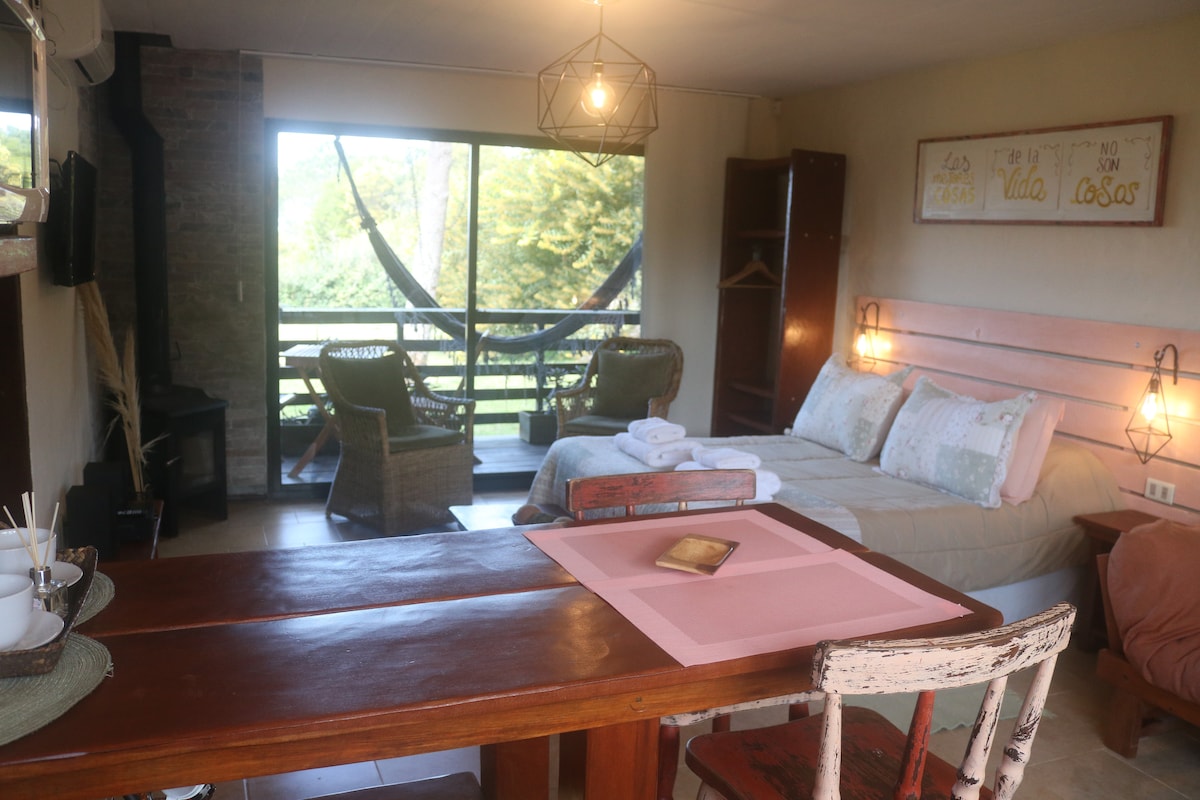 A spacious living area showcases a bed with soft linens, complemented by a dining table and chairs set nearby. A sliding glass door opens to a deck with hammocks, allowing natural light and views of the surrounding greenery to fill the space.