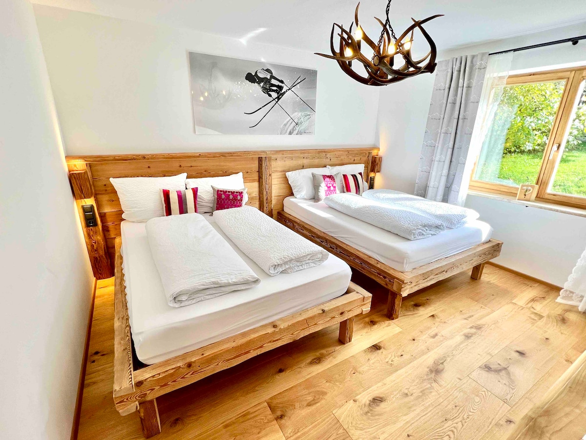 A serene bedroom is furnished with two wooden beds featuring built-in nightstands. Crisp linens are neatly arranged on each bed, complemented by decorative pillows. A large window offers natural light, while a piece of art depicting a skier adorns the wall, enhancing the alpine theme.