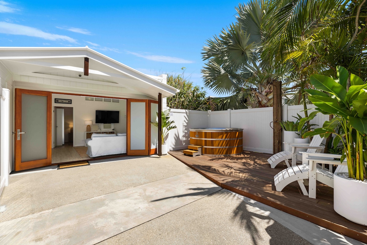 An inviting outdoor space features a wooden deck with a spa and white adirondack chairs surrounded by lush greenery. The cottage’s glass doors provide a view of the bedroom area, enhancing the connection to nature and outdoor living.