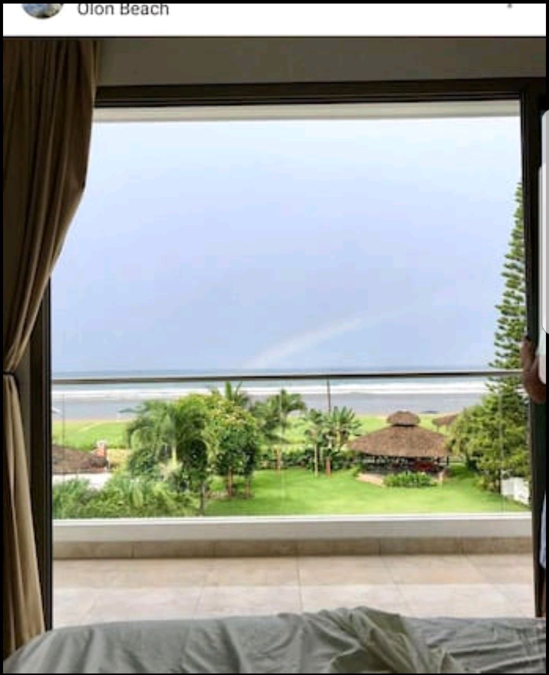 A spacious glass balcony offers a view of the ocean, framed by lush greenery and a beachfront gazebo. The horizon may display a subtle rainbow amidst the cloudy sky, while the interior hints at a comfortable bedding arrangement.