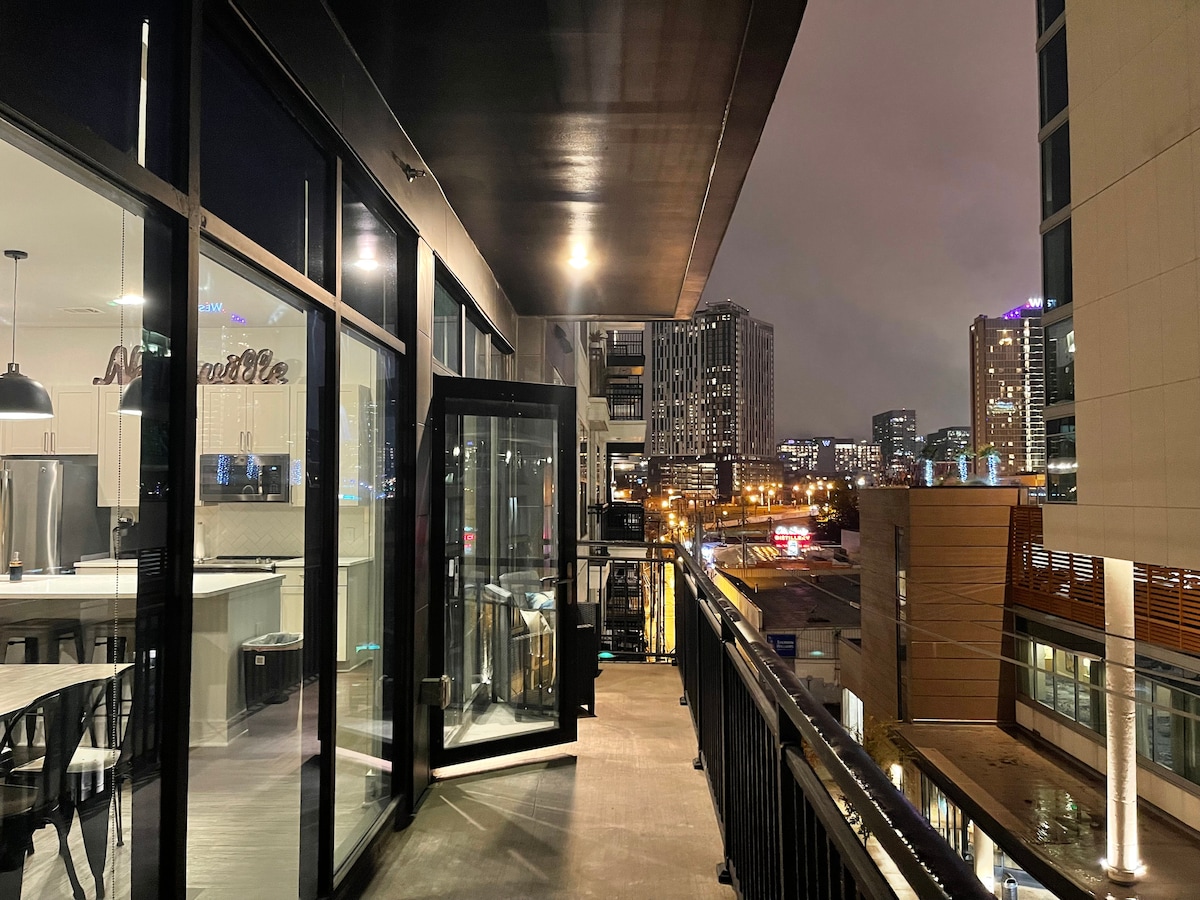 A private balcony extends from the corner unit, featuring modern railings and ample space for seating. The city skyline is visible in the background, illuminated by city lights at night. Glass doors lead into the living area, enhancing the open feel.