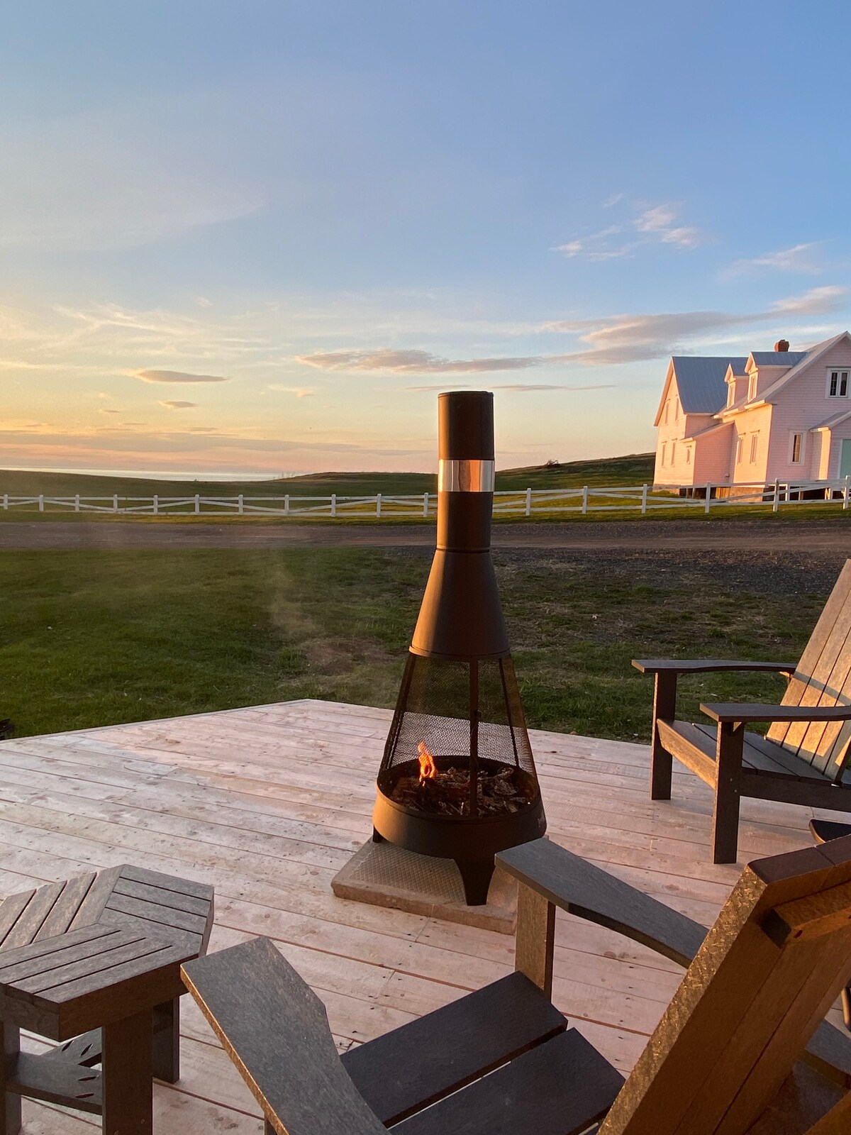 A sleek outdoor fireplace stands on a wooden deck, surrounded by dark wooden chairs. The sunset casts a warm glow over the landscape, with rolling green fields and a white house visible in the background.