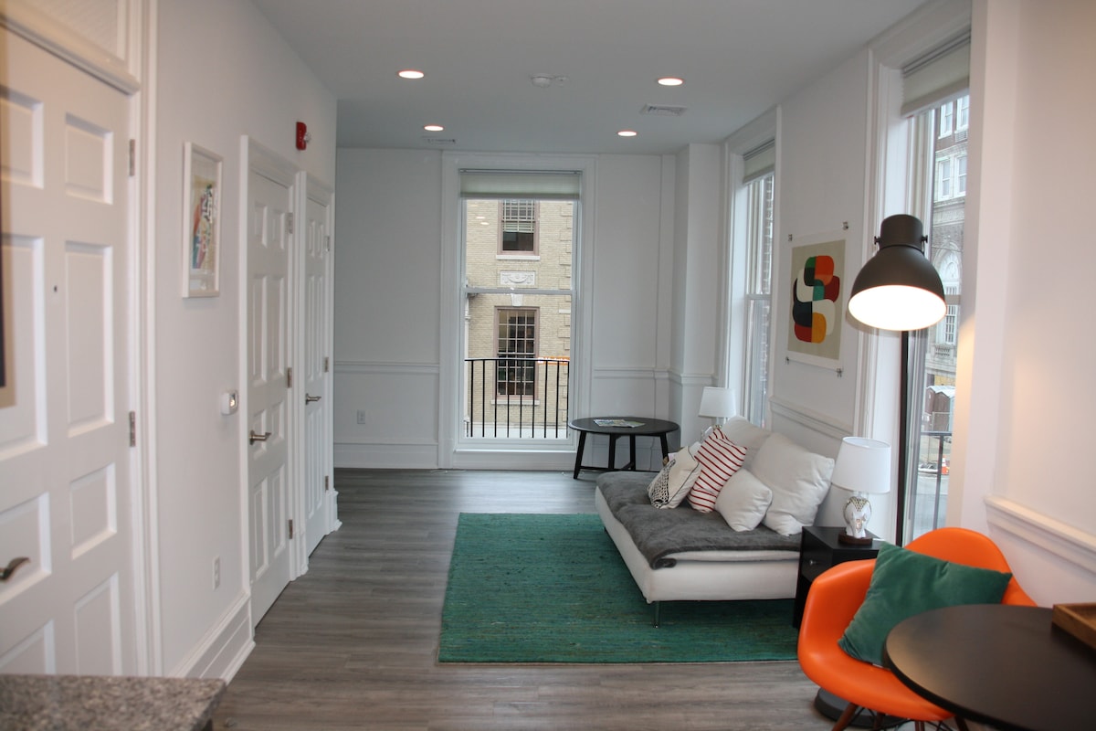 A modern living area is featured with a gray sofa adorned with colorful throw pillows. A blue area rug lies on the floor, complementing the light wood tones. Large windows allow natural light to fill the space, and a small round table with orange chairs is visible.
