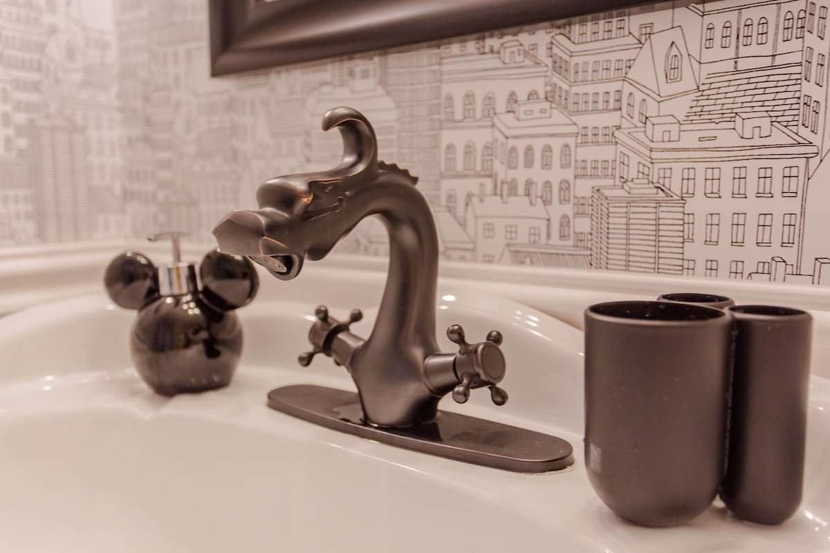 A unique bathroom sink features a dragon-shaped faucet in matte black. Nearby, a charming Mickey Mouse-shaped soap dispenser complements sleek black cups for toiletries, all set against a backdrop of monochrome cityscape wallpaper.