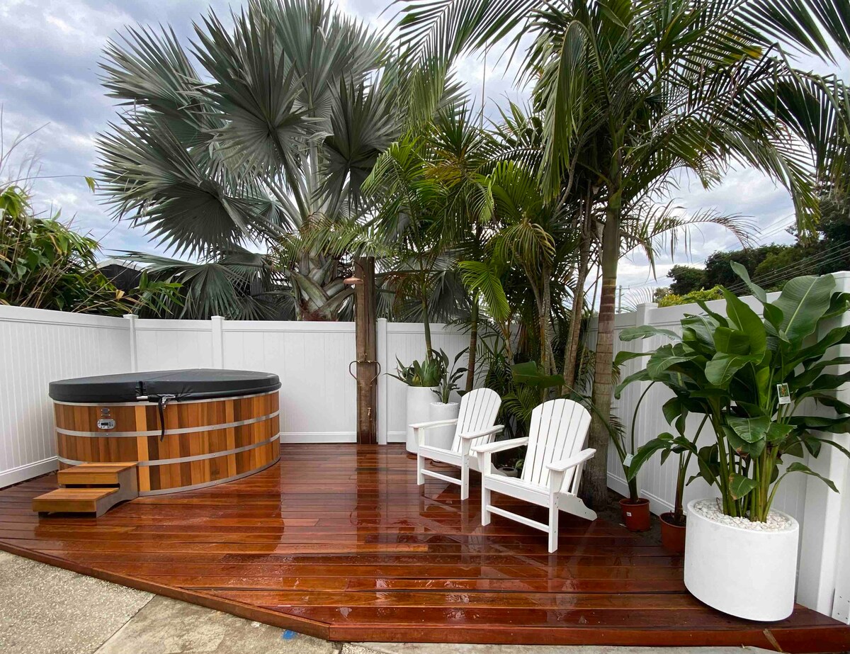 A private outdoor deck is presented with a wooden surface, featuring a circular hot tub surrounded by lush greenery. Two white Adirondack chairs are positioned alongside potted plants, creating a tranquil space for relaxation. A white fence encloses the area, enhancing privacy.