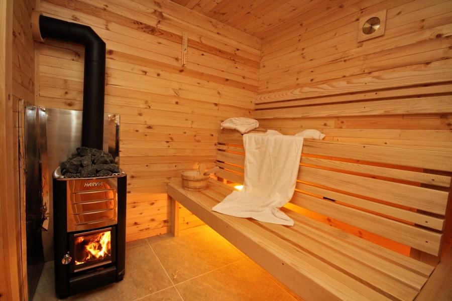 A welcoming sauna space is lined with warm wooden panels, featuring a stone heater emitting gentle warmth. Two benches offer seating, with soft towels draped thoughtfully. Subtle lighting enhances the cozy ambiance, creating a relaxing retreat.