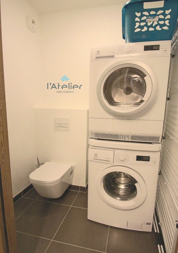 A compact laundry area features a stacked washer and dryer, both in white. A small basket is placed on top of the dryer. To the side, a modern toilet is visible, set against neutral-colored walls and tiled flooring, optimizing the space.