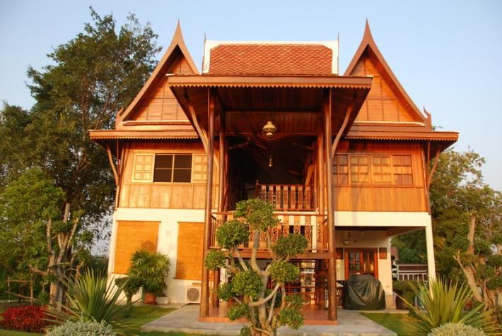 A traditional Thai-style house features distinctive peaked roofs and expansive wooden elements. The front porch is highlighted by decorative columns and surrounded by well-maintained greenery. Sunlight reflects off the wooden surfaces, enhancing the warm tones of the structure. A barbecue grill is discreetly placed nearby.