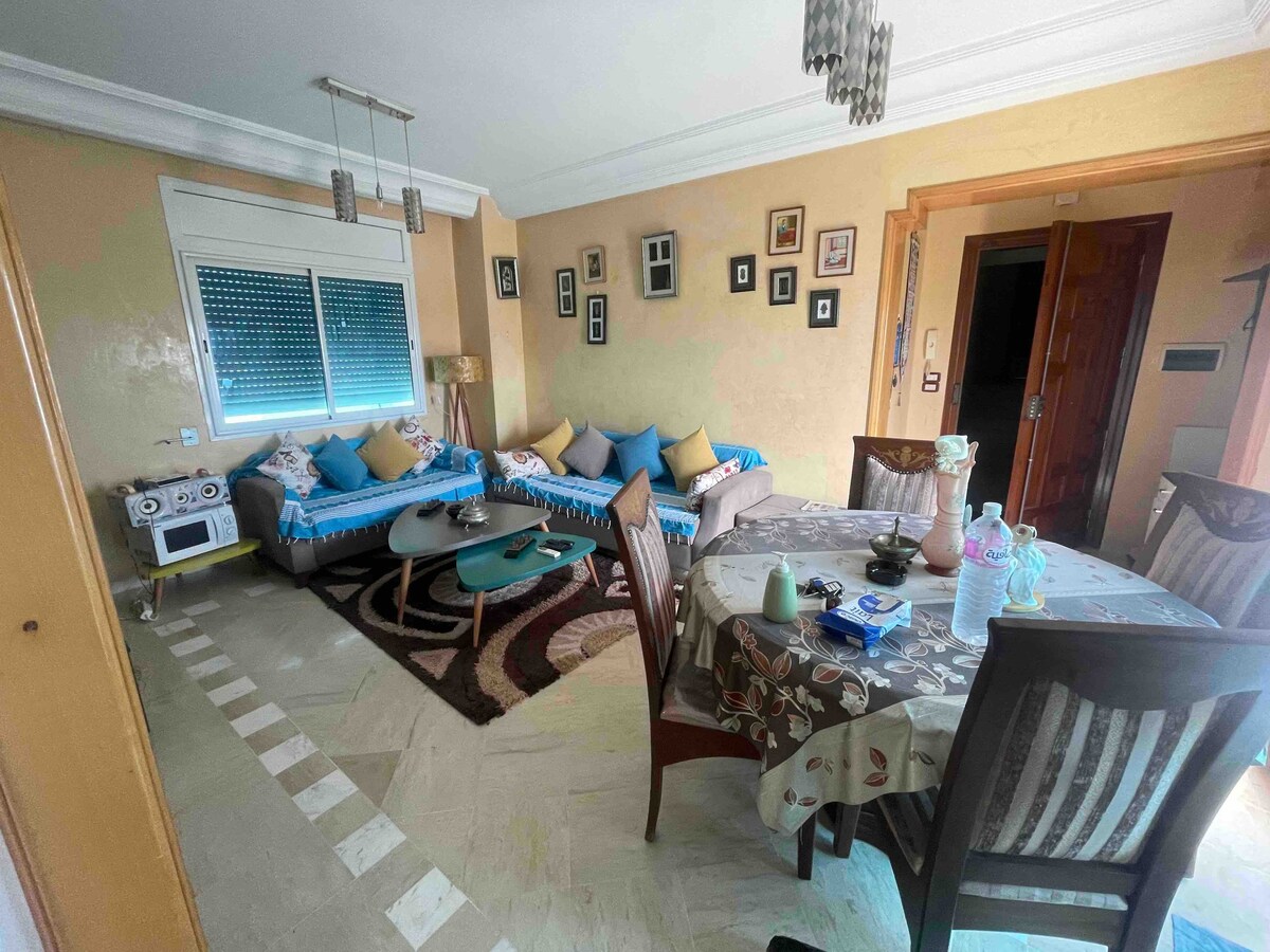 An open living and dining area is featured, showcasing two sofas adorned with cushions and a round glass coffee table at the center. A wooden dining table is set with patterned tableware. The walls display framed pictures, and natural light enters through a window with decorative blinds.